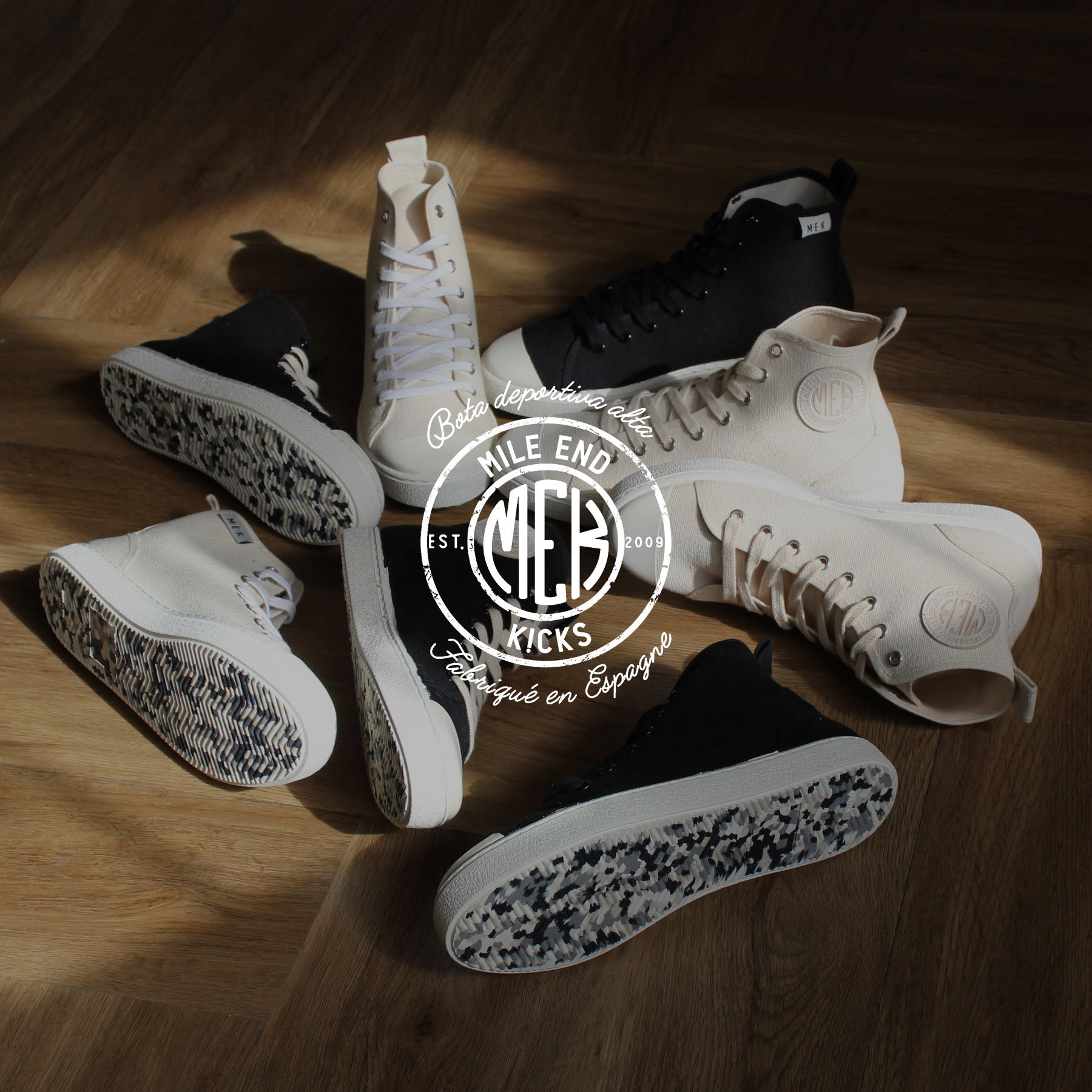 A mix of black and white high top canvas shoes on the floor. Light from a window shines over the scene. There is a logo overlaid on the image.