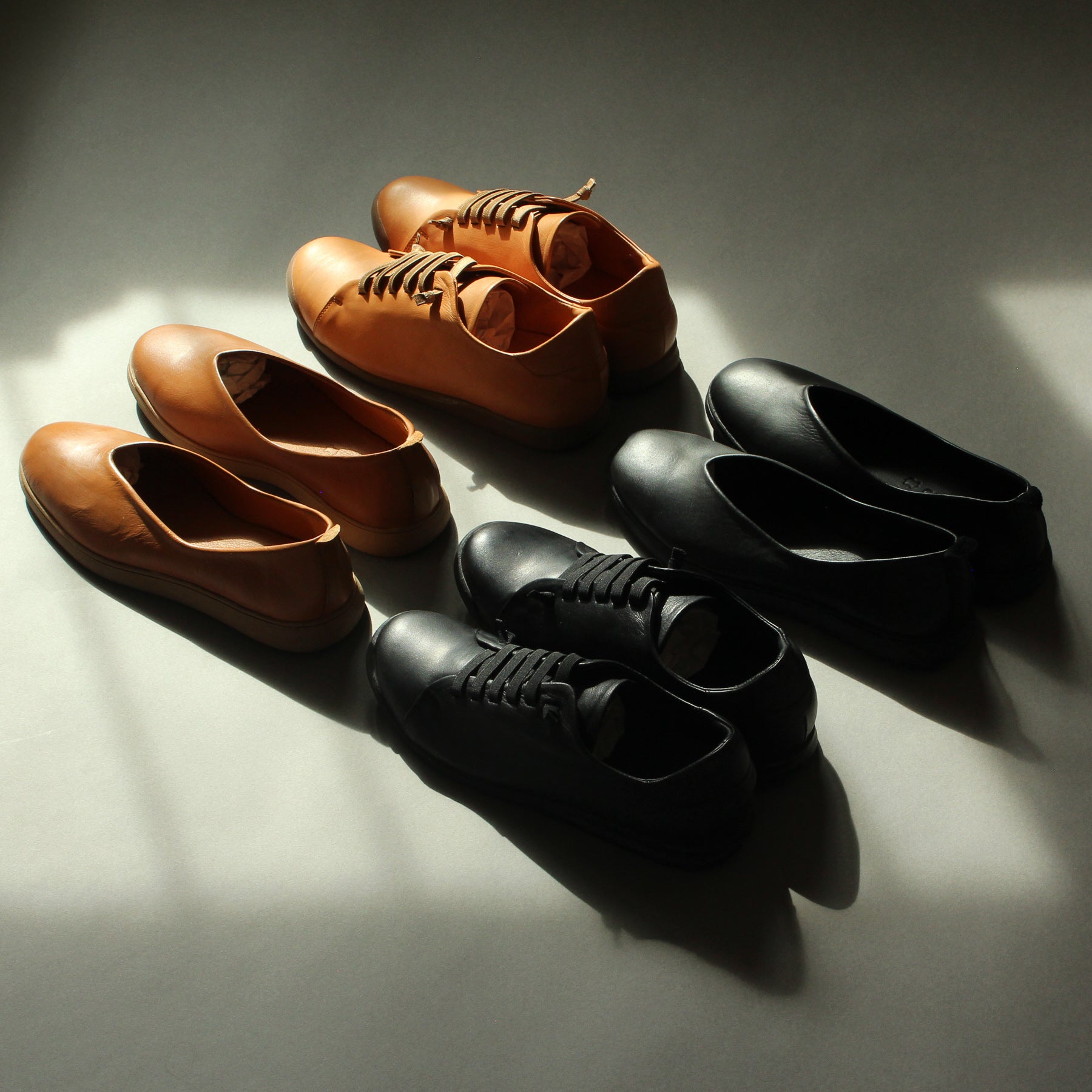 Different models of black and brown shoes are arranged in a diagonal pattern on a grey background. There is speckled light cast on the shoes and the backdrop creating contrast in the image.