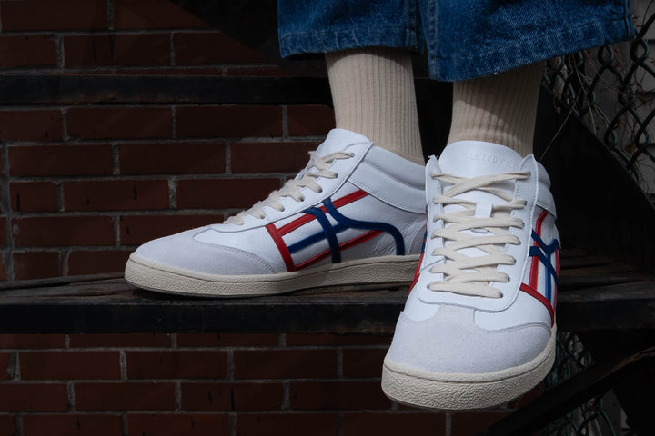 White sneakers with red and blue accents worn by a person standing in front of a brick wall.